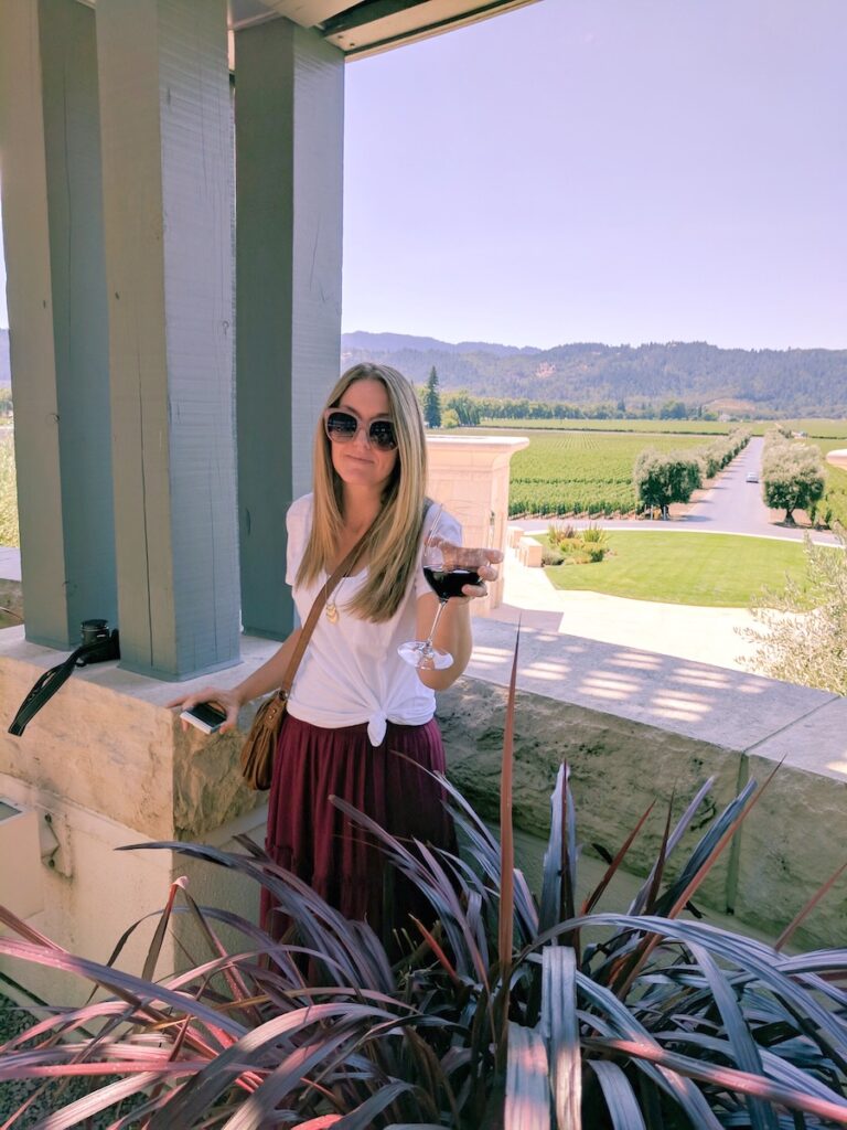 All about Travel Stories, a little blog about where we're going and where we've been. 
This is a photo of a blond woman wearing a white shirt and burgandy skirt,  holding a large glass of red wine, with a view of a Napa vineyard behind her.